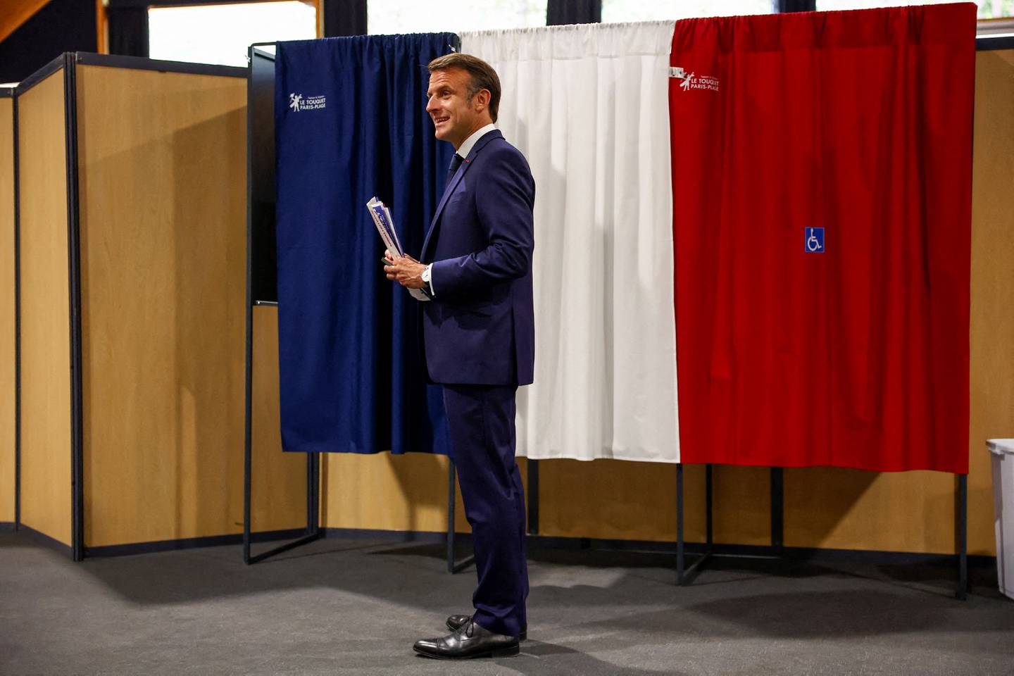 El presidente de Francia, Emmanuel Macron, se encuentra frente a una cabina electoral, adornada con cortinas que muestran los colores de la bandera de Francia, antes de emitir su voto para las elecciones al Parlamento Europeo.