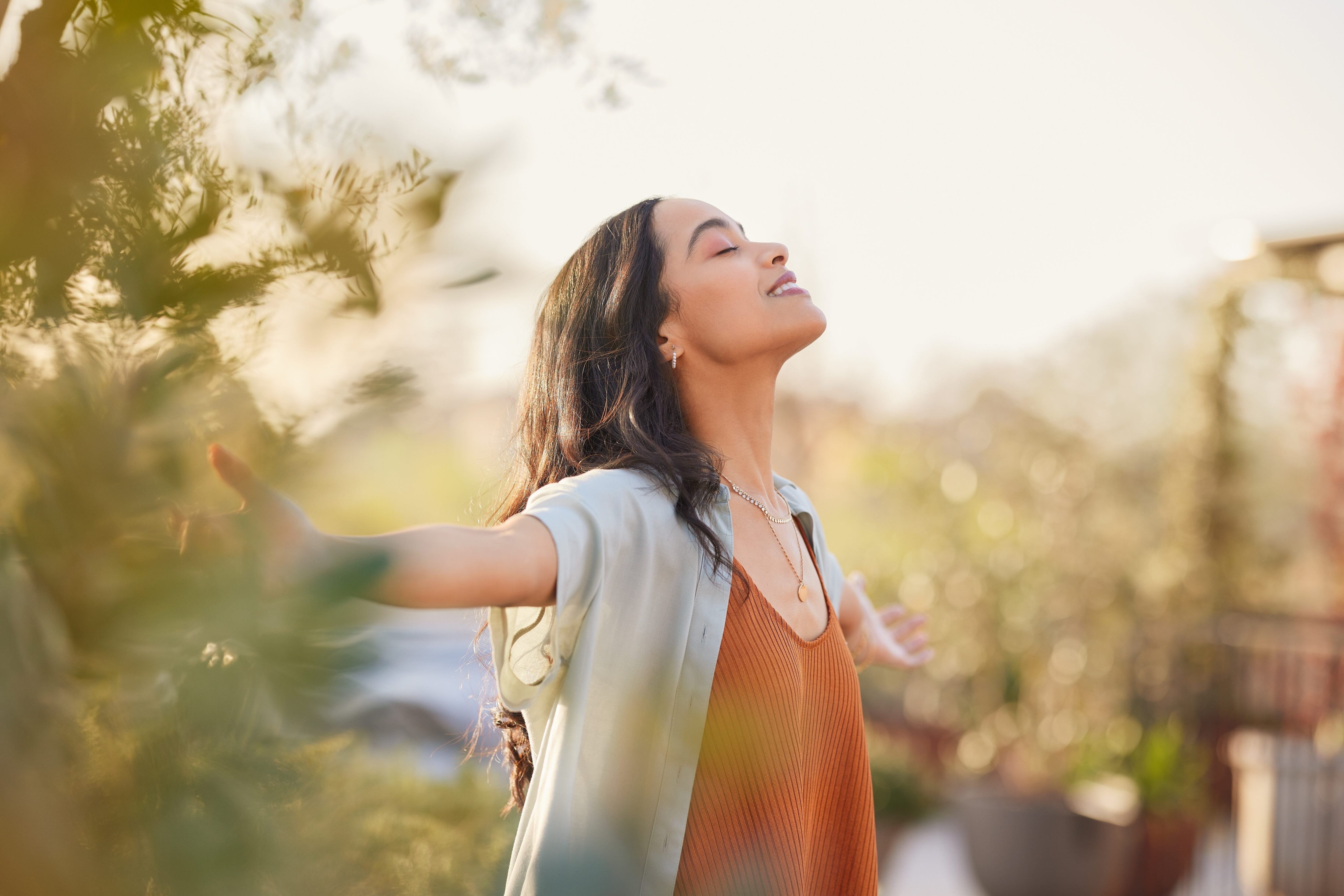 Young latin woman with arms outstretched breathing in fresh air during sunrise at the balcony. Girl enjoying nature while meditating during morning with open arms and closed eyes. Mindful woman relax.